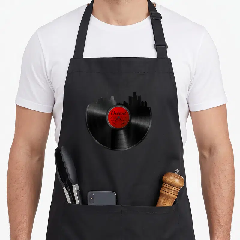 Tablier de cuisine noir avec illustration d'un vinyle noir dont le haut forme un skyline urbain au-dessus d'une étiquette rouge centrale.