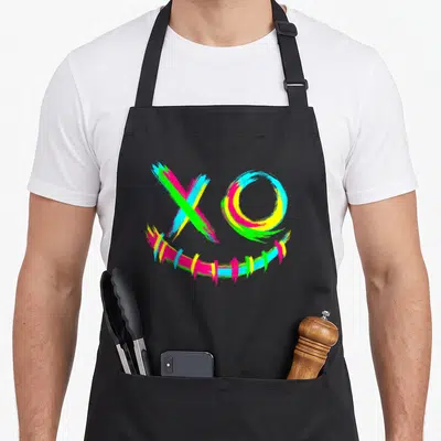 Tablier de cuisine noir avec un visage rave aux couleurs vives formé par des traits de pinceau multicolores.