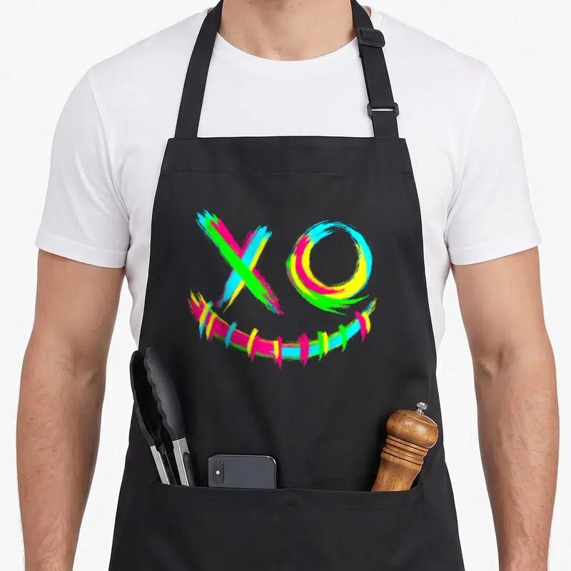 Tablier de cuisine noir avec un visage rave aux couleurs vives formé par des traits de pinceau multicolores.