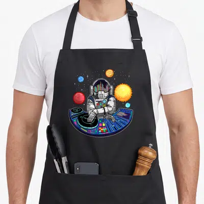 Tablier de cuisine noir avec illustration d'un astronaute DJ mixant sur une console colorée entourée de planètes spatiales.