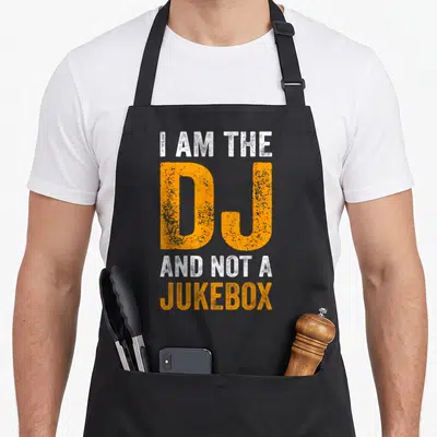 Tablier de cuisine noir avec l'inscription blanche et orange I Am The DJ And Not A Jukebox en typographie délavée.