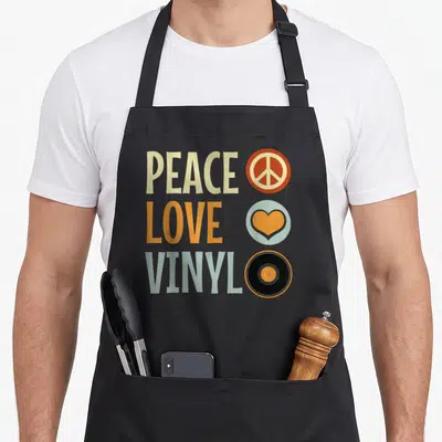 Tablier de cuisine noir avec Peace Love Vinyl, un disque vinyle, une paix et un cœur dans un style rétro rock vintage coloré.