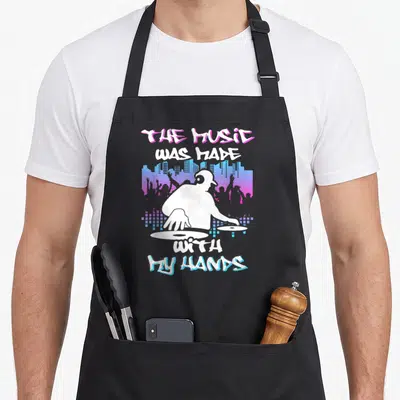 Tablier de cuisine noir avec illustration d'un DJ blanc et le texte The Music Was Made With My Hands.