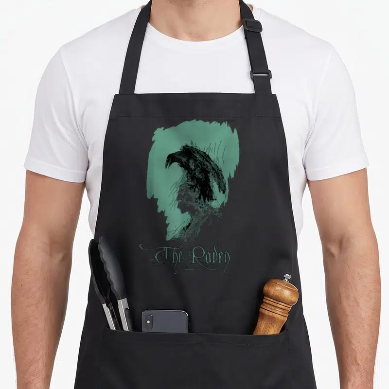 Tablier de cuisine noir avec illustration vintage d'un corbeau sur profil féminin et texte The Raven en typographie ornementale verte.