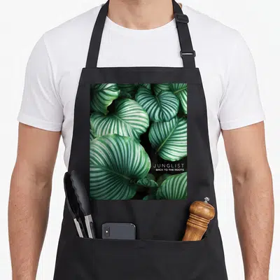 Tablier de cuisine noir avec motif de grandes feuilles vertes striées de blanc et le texte Junglist Back to the Roots en bas à droite.