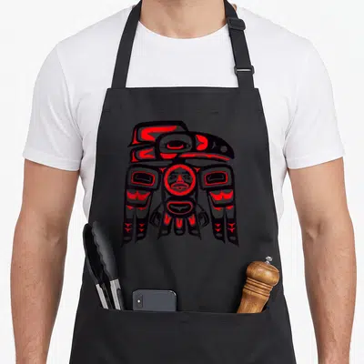 Tablier de cuisine noir avec un grand motif de totem corbeau aux couleurs rouge et gris foncé sur le devant.