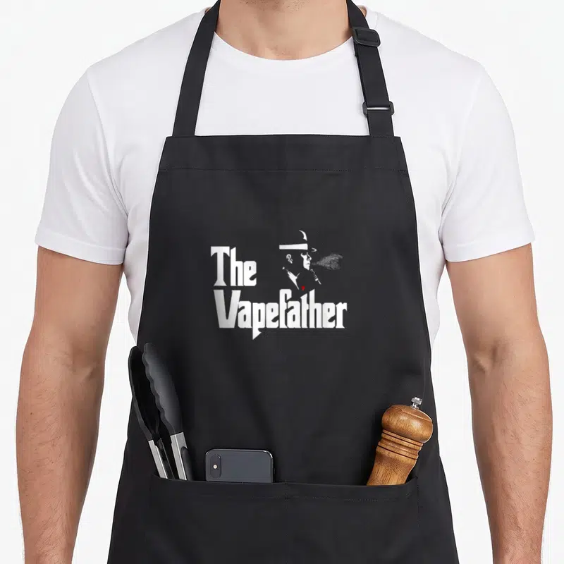 Tablier de cuisine noir avec une silhouette blanche fumant et le texte The Vapefather imprimé au centre.