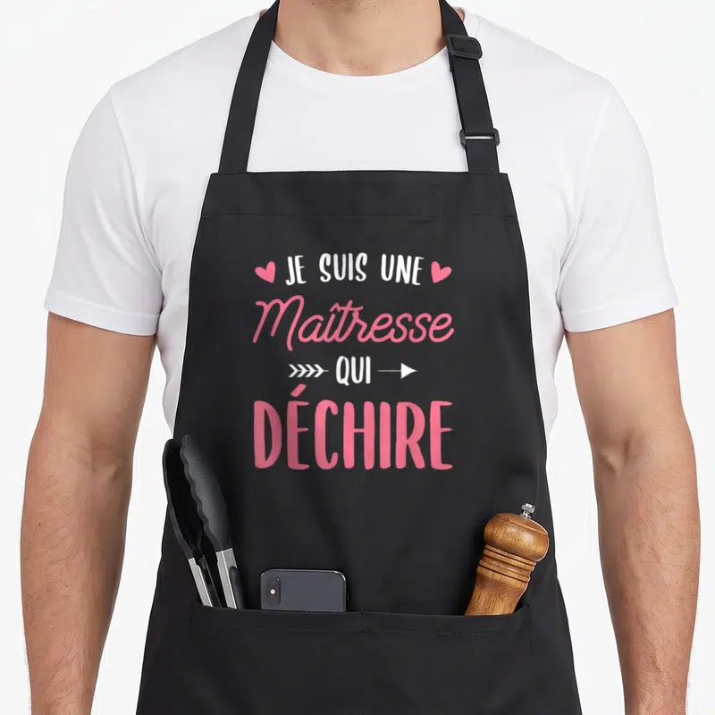 Tablier de cuisine noir avec le texte Je Suis Une Maîtresse Qui Déchire en rose et blanc, orné de petits cœurs et d'une flèche.