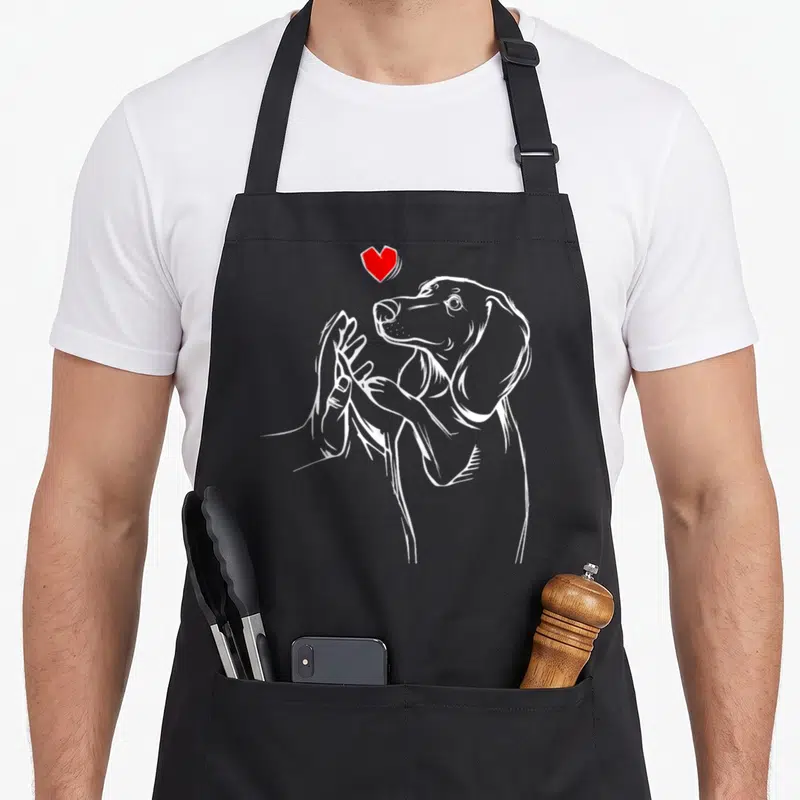 Tablier de cuisine noir avec illustration blanche d'un teckel et une main, surmonté d'un petit cœur rouge vif.