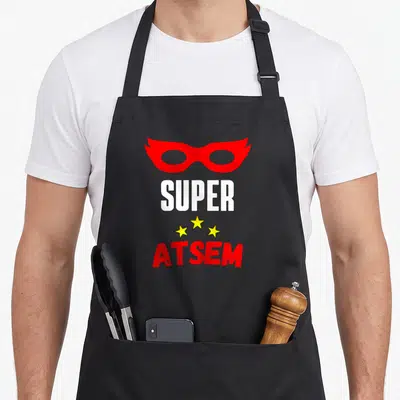 Tablier de cuisine noir avec un masque de super-héros rouge, le texte SUPER ATSEM et trois étoiles jaunes stylisées au centre.