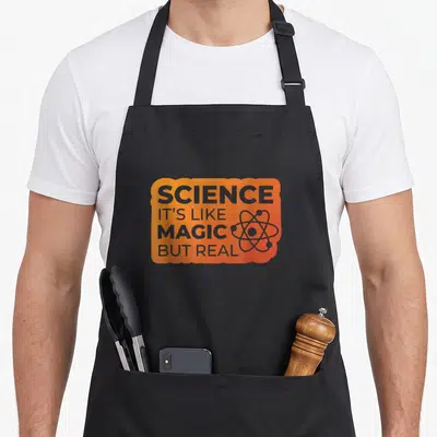 Tablier de cuisine noir avec une étiquette orange portant le texte Science it's like magic but real et un atome.