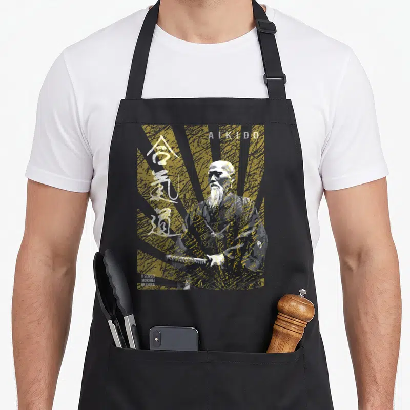Tablier de cuisine noir avec illustration stylisée du Maître d'Aïkido Morihei Ueshiba tenant un sabre sous des rayons dorés.