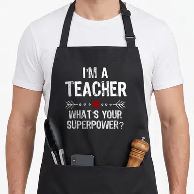 Tablier de cuisine noir avec le texte I'm a teacher What's your superpower et un cœur rouge entre deux flèches blanches.