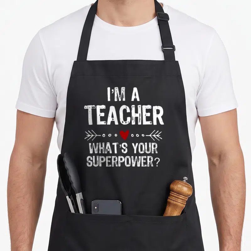 Tablier de cuisine noir avec le texte I'm a teacher What's your superpower et un cœur rouge entre deux flèches blanches.