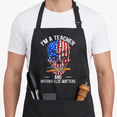 Tablier de cuisine noir avec un crâne aux couleurs du drapeau américain et des instruments scolaires dans la mâchoire.