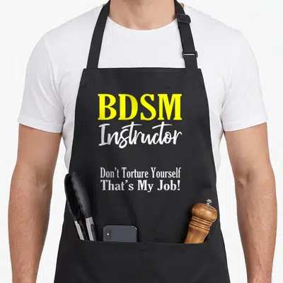 Tablier de cuisine noir avec l'inscription BDSM Instructor en jaune et blanc sur fond uni.