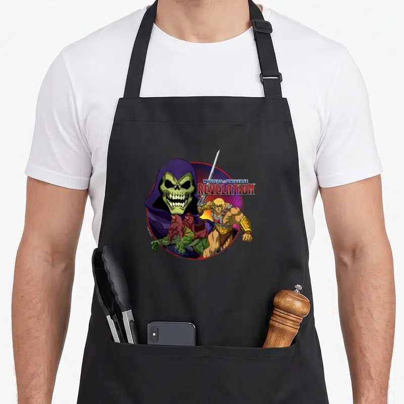Tablier de cuisine noir avec illustration circulaire de He-Man et Skeletor aux couleurs vives.