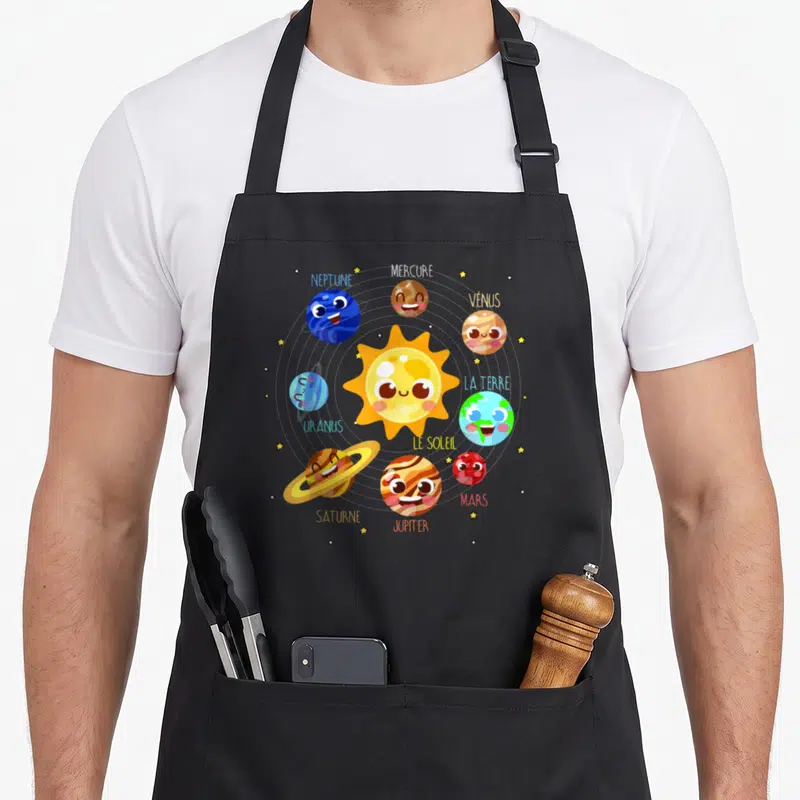 Tablier de cuisine noir avec un système solaire coloré montrant le Soleil et neuf planètes aux visages souriants et leurs noms respectifs.