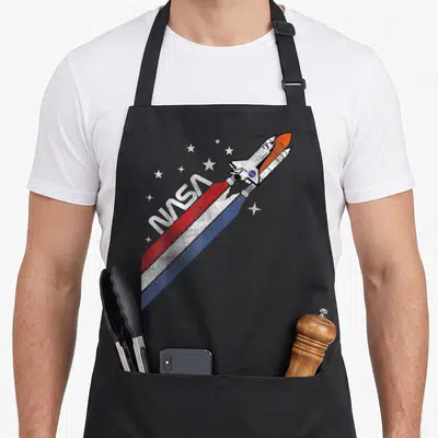 Tablier de cuisine noir avec une illustration de navette spatiale NASA et des étoiles sur fond uni.