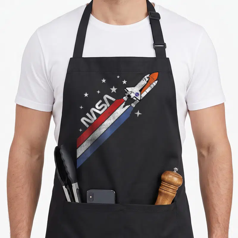 Tablier de cuisine noir avec une illustration de navette spatiale NASA et des étoiles sur fond uni.