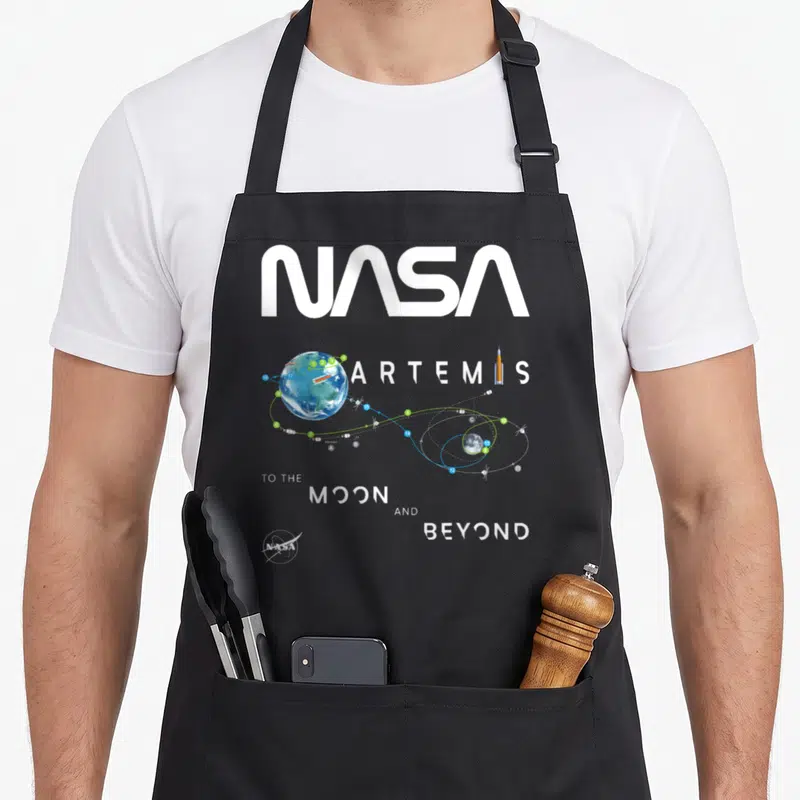 Tablier de cuisine noir avec l'insigne de mission Artemis, une carte orbitale et le texte To the Moon and Beyond en typographie blanche.