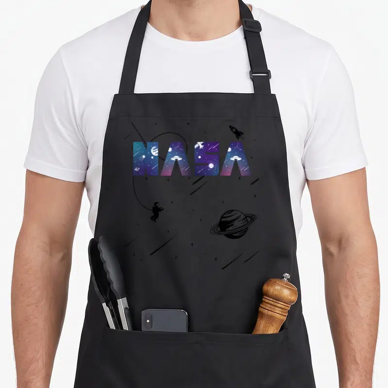 Tablier de cuisine noir avec le logo NASA coloré, un astronaute, des planètes et des engins volants non identifiés imprimés sur le devant.