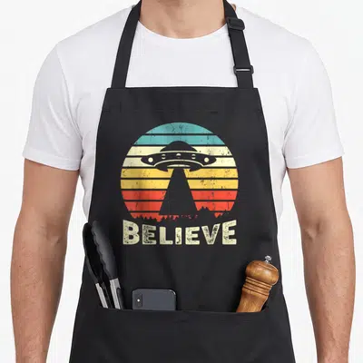 Tablier de cuisine noir avec illustration retro d'un vaisseau spatial et le mot BELIEVE en bas.