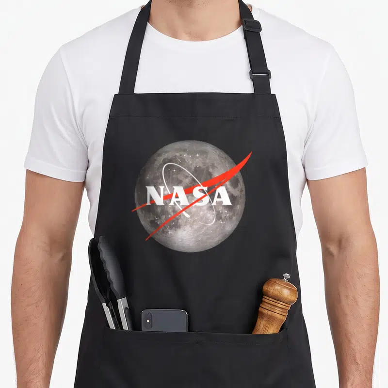 Tablier de cuisine noir avec une lune texturée et le logo NASA traversé par une ligne rouge.