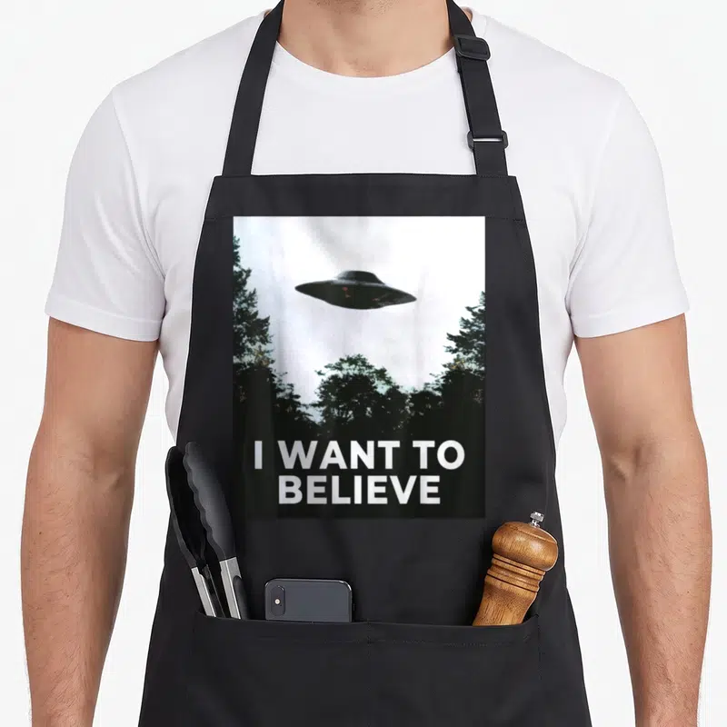 Tablier de cuisine noir avec un vaisseau spatial et l'inscription I WANT TO BELIEVE au-dessus d'arbres sombres.