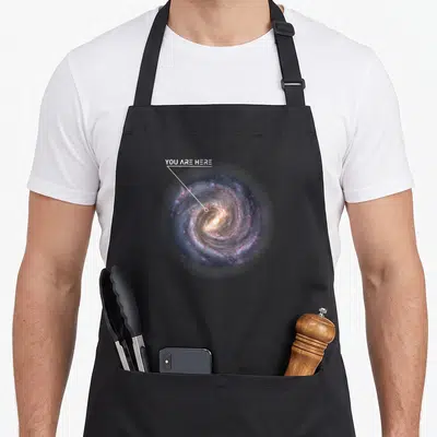 Tablier de cuisine noir avec illustration de galaxie spirale et texte You Are Here pointant vers le centre stellaire.