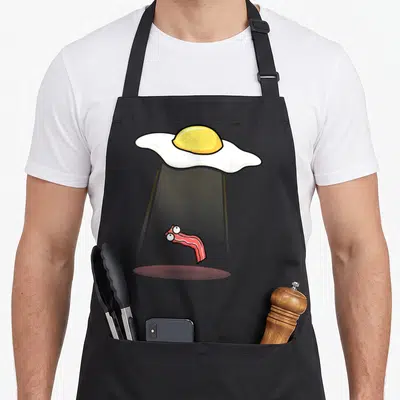 Tablier de cuisine noir avec illustration d'un oeuf ovni enlevant une lanière de bacon surprise.