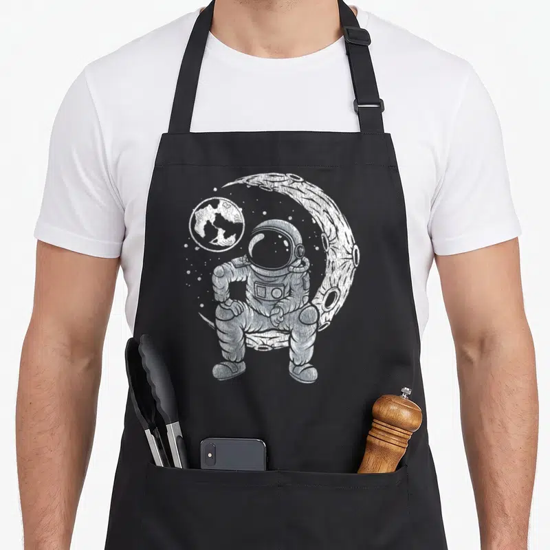 Tablier de cuisine noir avec illustration blanche d'un astronaute assis sur la Lune et la Terre en orbite.