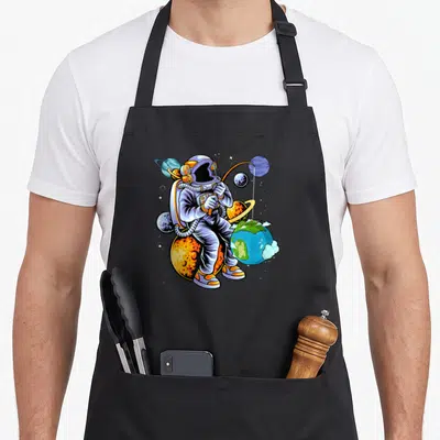 Tablier de cuisine noir avec illustration d'un astronaute pêchant depuis une planète orange au milieu de l'univers.