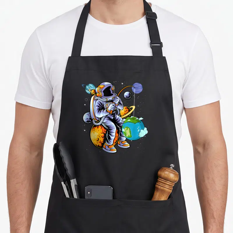 Tablier de cuisine noir avec illustration d'un astronaute pêchant depuis une planète orange au milieu de l'univers.