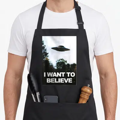 Tablier de cuisine noir avec un ufo flottant au-dessus d'arbres et le texte I WANT TO BELIEVE en blanc.