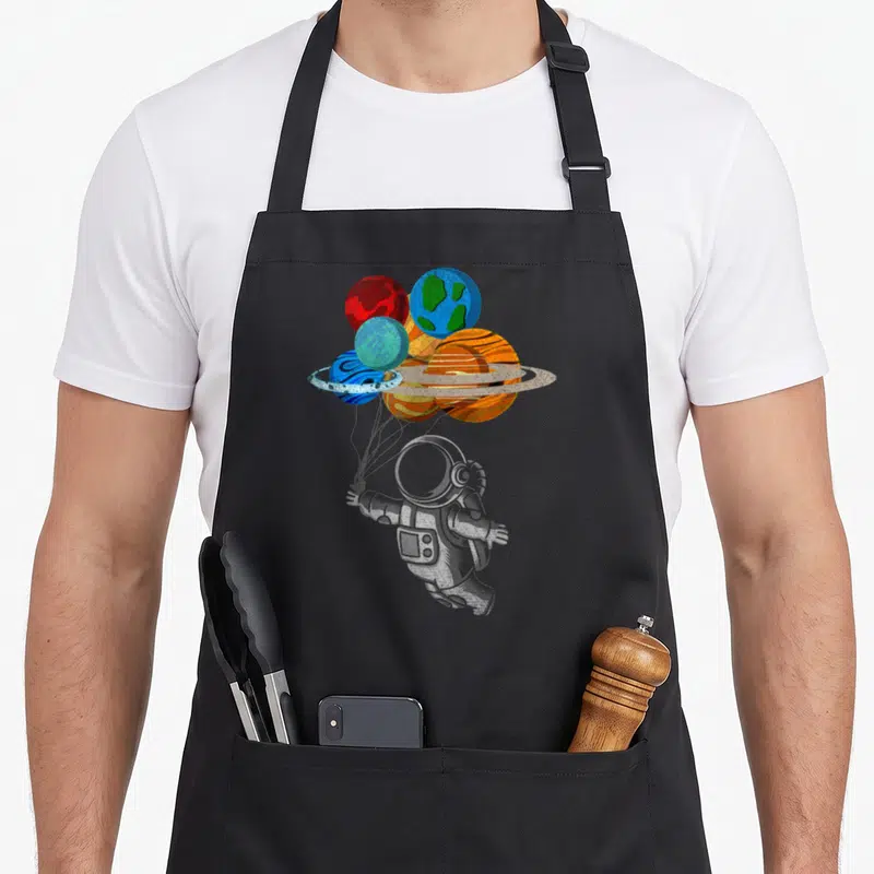 Tablier de cuisine noir avec un astronaute tenant des ballons représentant les planètes du système solaire en couleurs vives.