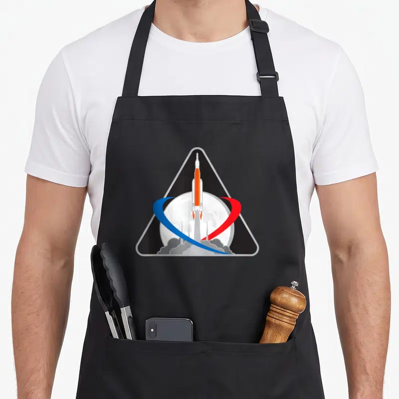 Tablier de cuisine noir avec le logo triangulaire du lanceur Orion SLS Artemis 1 devant la Lune et des orbites rouge et bleue.