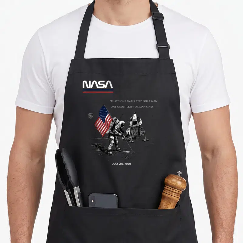 Tablier de cuisine noir avec illustration astronautique, drapeau américain et logo spatial en haut à gauche.