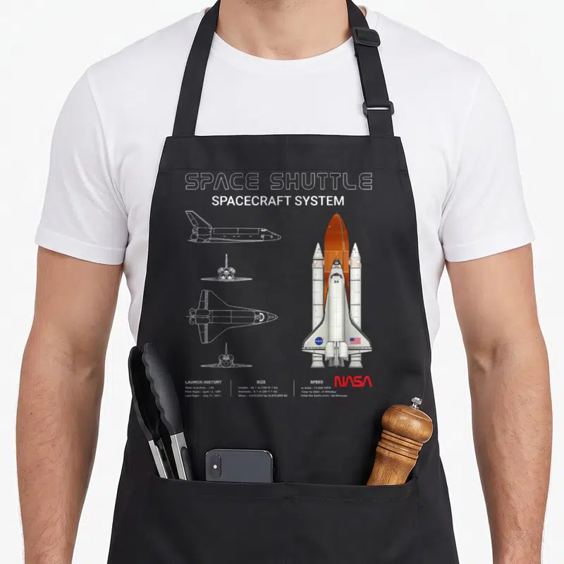 Tablier de cuisine noir avec illustration colorée d'un lanceur spatial et vues techniques en dessin au trait blanc sur fond gris.