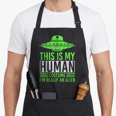 Tablier de cuisine noir avec un ovni vert et le texte This Is My Human Costume I'm Really An Alien imprimé au centre.