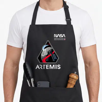 Tablier de cuisine noir avec le logo triangulaire de la mission Artemis NASA et une orbite rouge sur fond lunaire.