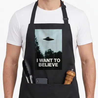Tablier de cuisine noir imprimé avec un engin volant au-dessus d'arbres et le texte I Want To Believe en bas.