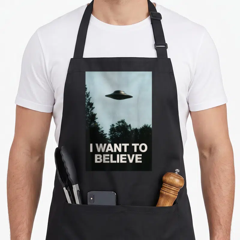 Tablier de cuisine noir imprimé avec un engin volant au-dessus d'arbres et le texte I Want To Believe en bas.