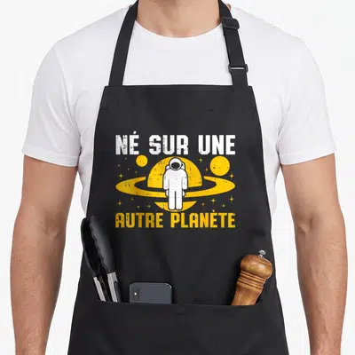 Tablier de cuisine noir avec illustration d'astronaute blanc devant une planète jaune et texte humoristique.
