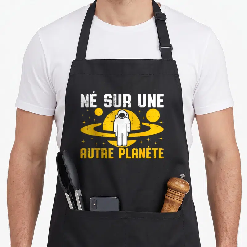 Tablier de cuisine noir avec illustration d'astronaute blanc devant une planète jaune et texte humoristique.