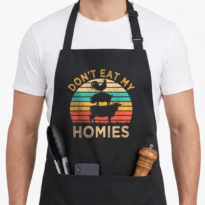 Tablier de cuisine noir avec illustration vintage montrant un coq, un porc et un bœuf sur fond de soleil couchant coloré.