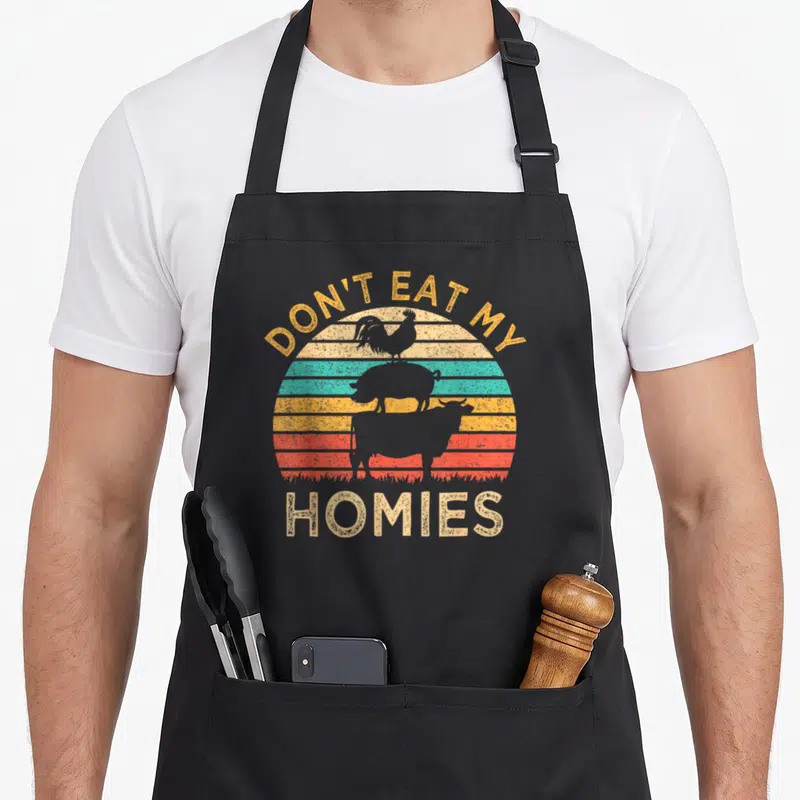 Tablier de cuisine noir avec illustration vintage montrant un coq, un porc et un bœuf sur fond de soleil couchant coloré.