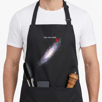 Tablier de cuisine noir avec une illustration de galaxie rose et bleue marquée par un repère rouge You Are Here.
