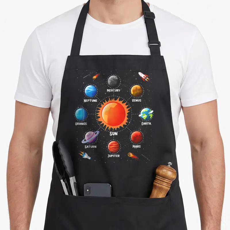 Tablier de cuisine noir avec illustration colorée du système solaire incluant le soleil, les planètes et des fusées spatiales.