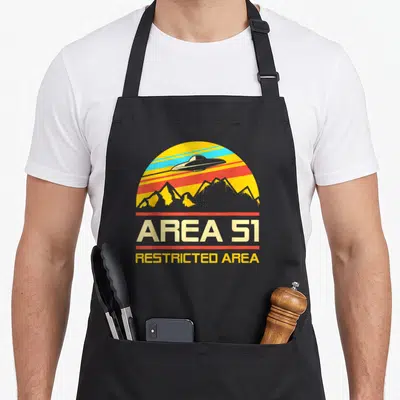 Tablier de cuisine noir avec logo Area 51 rétro montrant un vaisseau spatial au-dessus de montagnes aux couleurs vives.